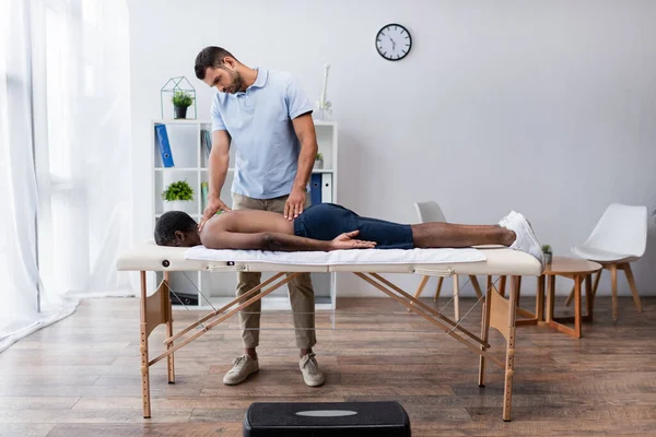 Full length view of young rehabilitologist near african american man lying face down on massage table — Stock Photo
