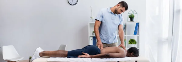 Homme afro-américain allongé sur une table de massage près du médecin dans un centre de réadaptation, bannière — Photo de stock
