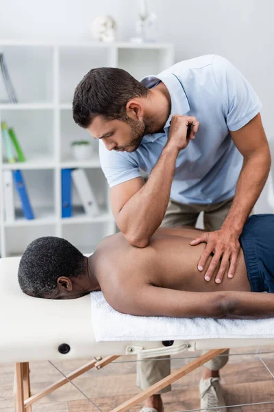 Hombre afroamericano acostado en la mesa de masaje durante la masoterapia en el centro de rehabilitación - foto de stock