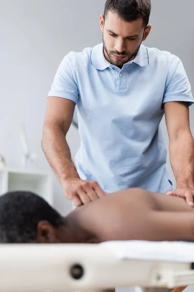 Joven quiropráctico haciendo masaje de espalda mientras trata borrosa hombre afroamericano - foto de stock