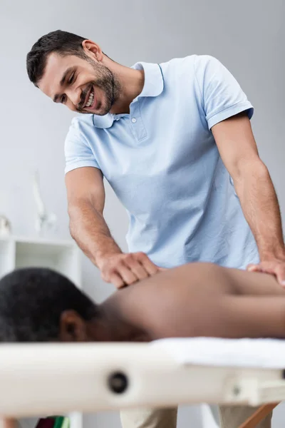 Sonriente masajista haciendo tratamiento masaje a borrosa afroamericano hombre en centro de rehabilitación - foto de stock