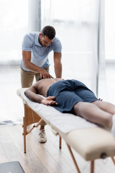 Joven fisioterapeuta haciendo masaje de cuello a hombre afroamericano en primer plano borroso - foto de stock