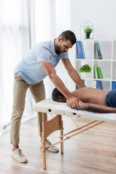 Young physiotherapist doing rehabilitation procedure to african american man on massage table — Stock Photo