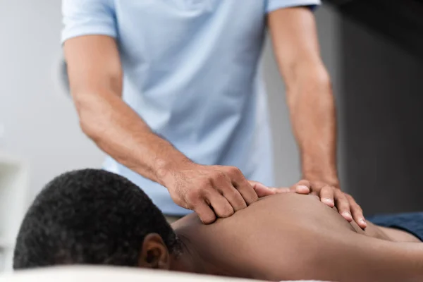 Fisioterapeuta borrosa haciendo masaje de espalda al hombre afroamericano durante el tratamiento de rehabilitación - foto de stock