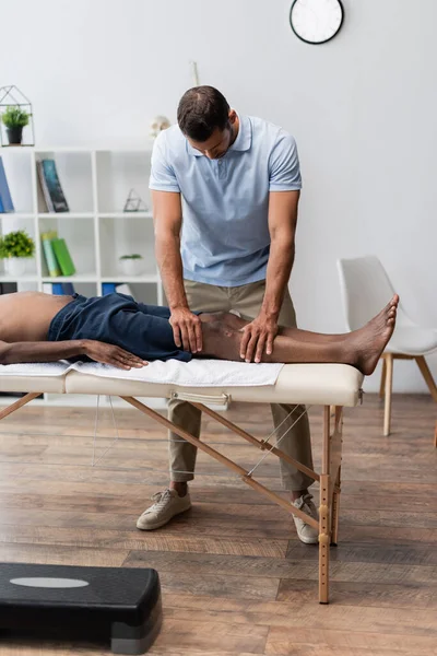 Chiropractor massaging knee of african american man in rehabilitation center — Stock Photo