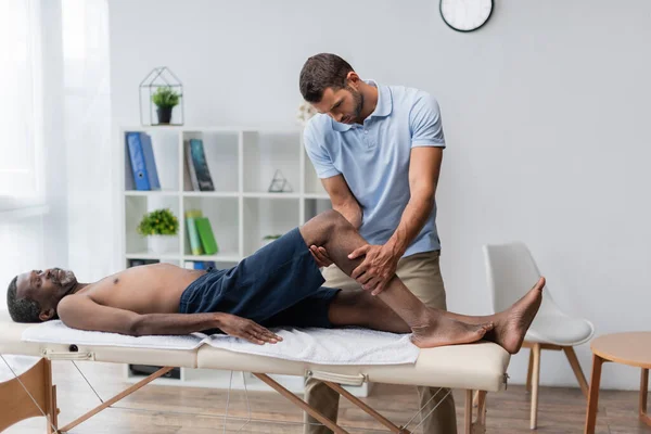 Rehabilitologist treating middle aged african american man lying on massage table — Fotografia de Stock