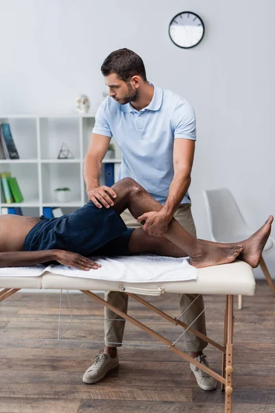 Rehabilitólogo tratamiento afroamericano hombre acostado en mesa de masaje - foto de stock