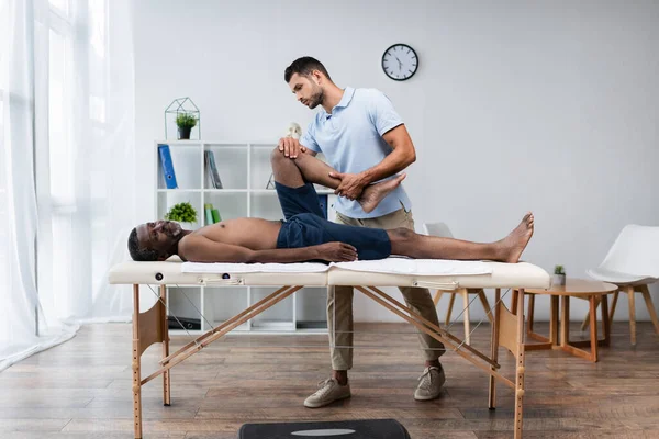 Full length view of rehabilitologist treating african american man on massage table — Stock Photo