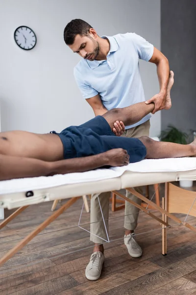 Quiropráctico masajeando pierna de hombre afroamericano en clínica de rehabilitación - foto de stock