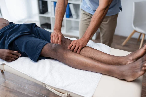 Vista recortada de quiropráctico masajeando rodilla de hombre afroamericano en centro de rehabilitación - foto de stock