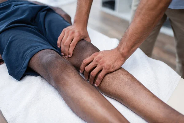 Vista recortada de masajista masajeando rodilla de hombre afroamericano en centro de rehabilitación - foto de stock