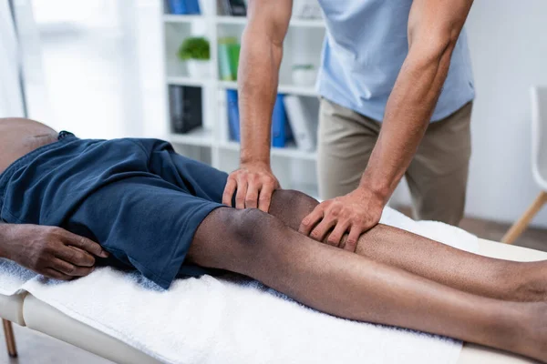 Partial view of rehabilitologist massaging leg of african american man — Foto stock
