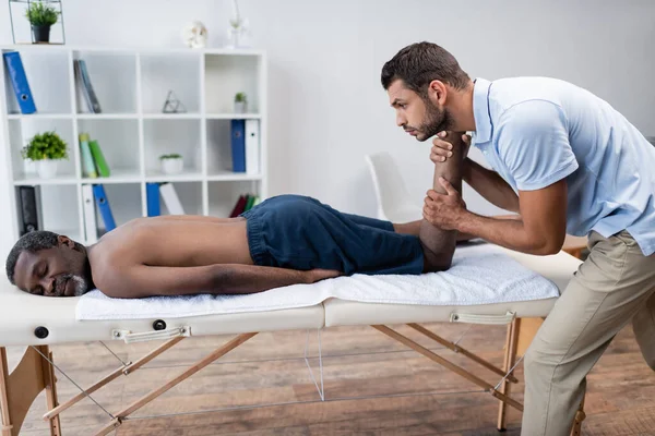 Homme mûr afro-américain couché sur la table de massage pendant la massothérapie dans le centre de réadaptation — Photo de stock