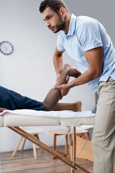 Jeune réhabilitologue étirement jambe du patient afro-américain sur table de massage — Photo de stock