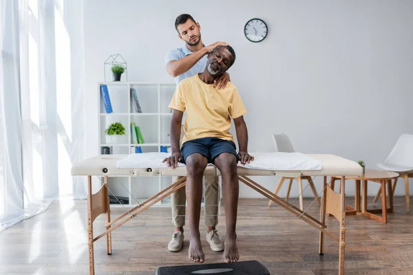 Doctor examining mature african american man sitting on massage table in rehabilitation center — Fotografia de Stock
