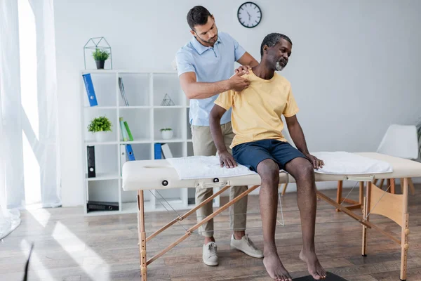 Fisioterapeuta examinando ombro de homem americano africano maduro sentado na mesa de massagem — Fotografia de Stock