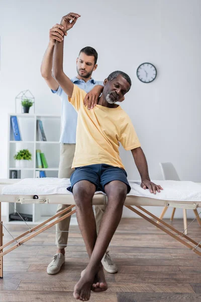 Physiotherapist raising hand of african american patient sitting on massage table — Stock Photo