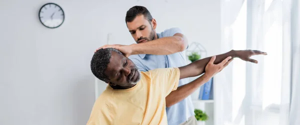 Physiotherapeut streckt Arm eines Afroamerikaners aus, während er ihn im Reha-Zentrum untersucht, Banner — Stockfoto