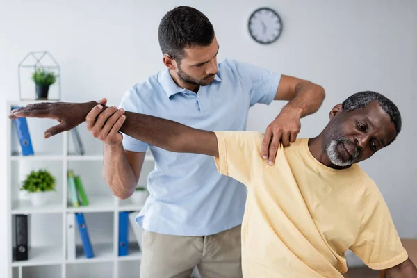 Fisioterapeuta examinando brazo y hombro de hombre afroamericano maduro en el hospital - foto de stock