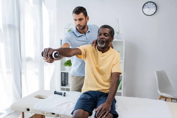 Physical therapist supporting african american man working out with dumbbell while sitting on massage table — Stock Photo