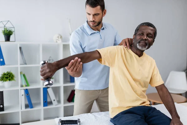 Hospital trainer assisting mature african american patient training with dumbbell — Foto stock