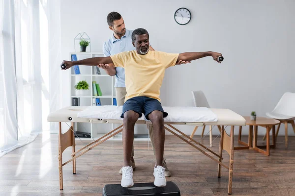Reabilitologista assistindo o homem afro-americano treinando com halteres na mesa de massagem — Fotografia de Stock