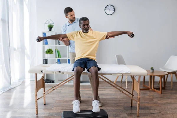 Rehabilitólogo asiste a afroamericano hombre entrenamiento con mancuernas en mesa de masaje - foto de stock