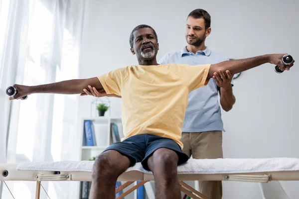 Physiotherapeut unterstützt reiferen afrikanisch-amerikanischen Mann beim Training mit Hanteln — Stockfoto