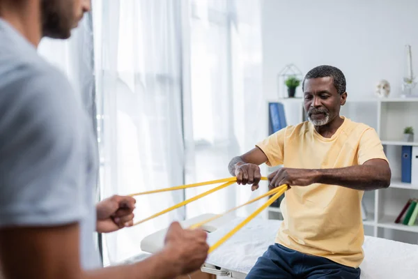 Reabilitologista desfocado e paciente afro-americano fazendo exercício com elásticos — Fotografia de Stock