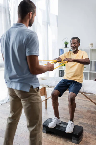 Hombre afroamericano haciendo ejercicio de rehabilitación con elásticos y fisioterapeuta borroso - foto de stock