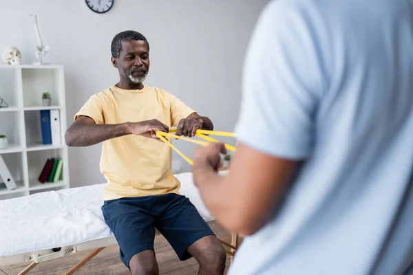 Mature african american man doing exercise with elastics together with blurred rehabilitologist — Stock Photo