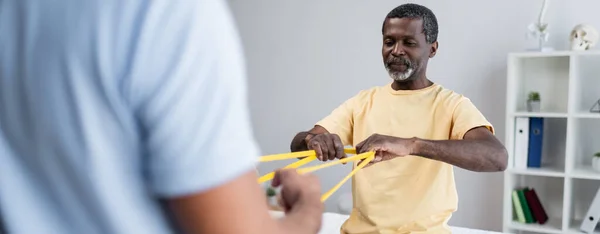 African american man working out with elastics together with blurred rehabilitologist, banner — Fotografia de Stock