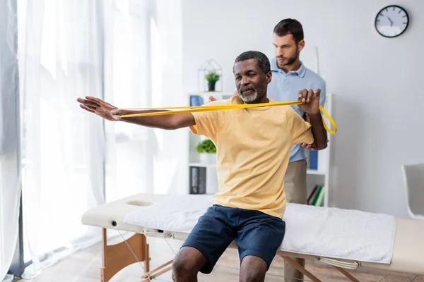 Homem americano africano de meia idade sentado na mesa de massagem e treinamento com elásticos perto de reabilitologista — Fotografia de Stock