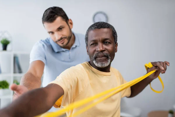 Maduro afroamericano hombre entrenamiento con elásticos cerca borrosa entrenador en centro de rehabilitación - foto de stock