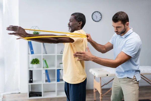 Joven fisioterapeuta asistiendo maduro afroamericano hombre entrenamiento con elásticos - foto de stock