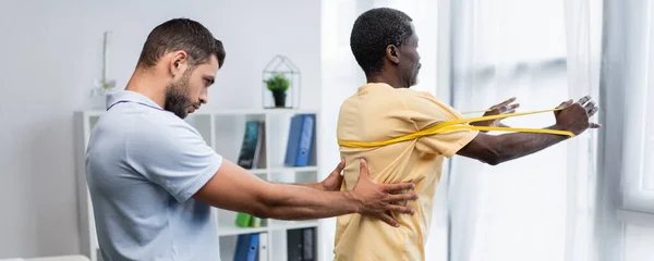 Physiotherapeut unterstützt afrikanisch-amerikanische Männer mittleren Alters beim Training mit Gummizügen, Banner — Stockfoto