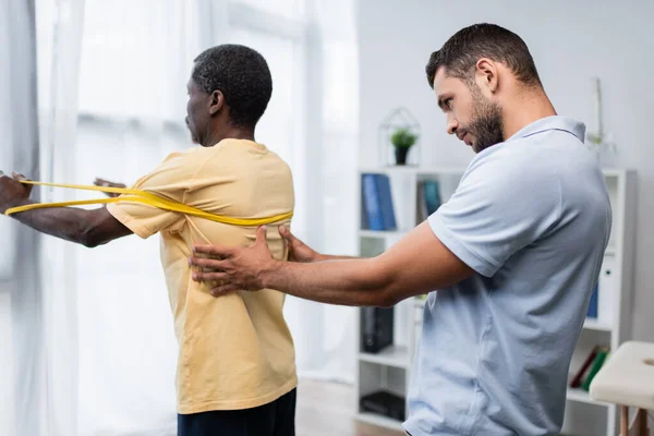 Entrenador ayudando a hombre afroamericano a hacer ejercicio con banda elástica en centro de rehabilitación - foto de stock