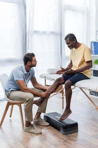 Reabilitologista tocando perna do homem afro-americano falando com ele enquanto sentado na mesa de massagem — Fotografia de Stock