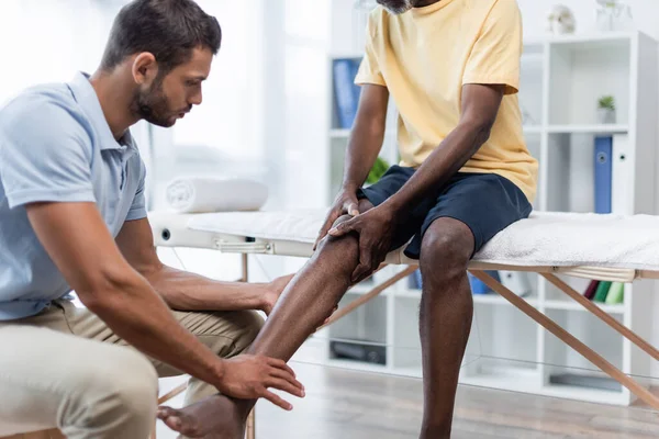 Young rehabilitologist touching leg of african american man sitting on massage table — Stockfoto