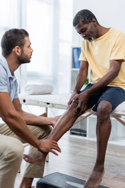 Hombre afroamericano maduro tocando la rodilla mientras habla con el médico en el centro de rehabilitación - foto de stock