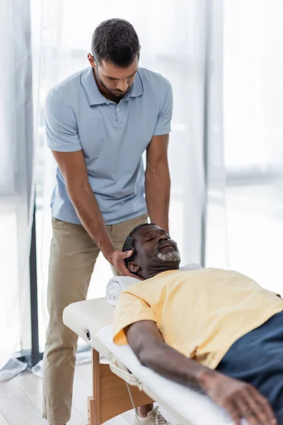 Joven fisioterapeuta tratamiento maduro africano americano hombre en mesa de masaje - foto de stock