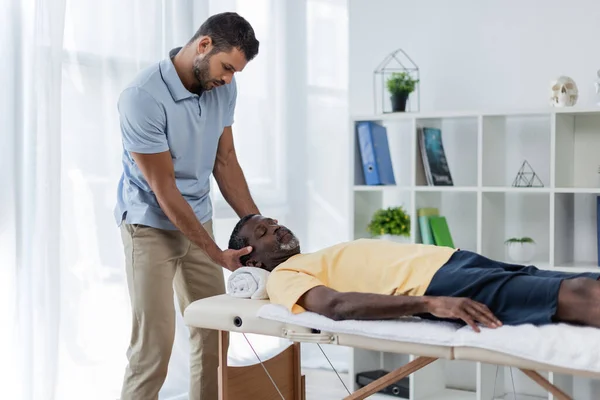 Uomo afroamericano con gli occhi chiusi sdraiato sul lettino da massaggio durante il trattamento di riabilitazione — Foto stock