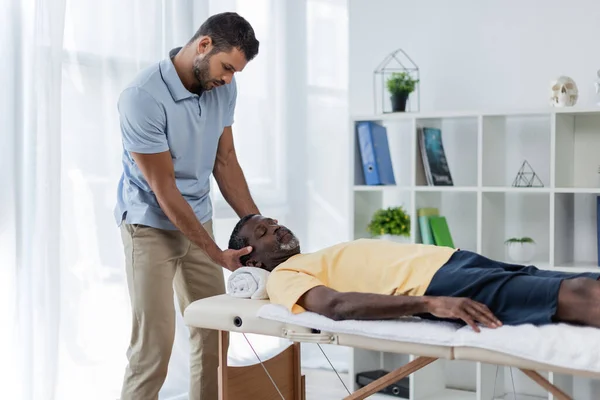 Hombre afroamericano con los ojos cerrados acostado en la mesa de masaje durante el tratamiento de rehabilitación - foto de stock