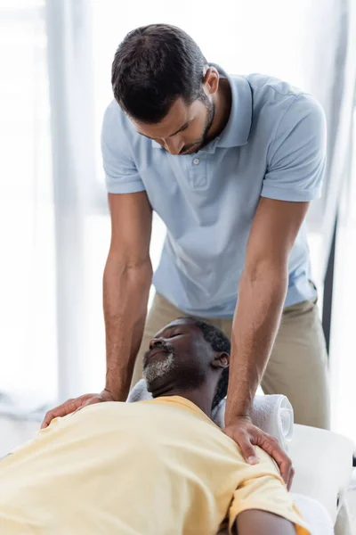 Hombre afroamericano maduro acostado en la mesa de masaje durante el tratamiento de rehabilitación - foto de stock
