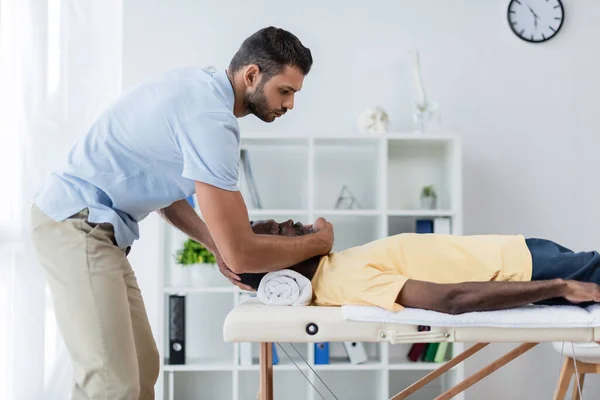 Vista lateral de un joven fisioterapeuta que trata a un paciente afroamericano en un centro de rehabilitación - foto de stock