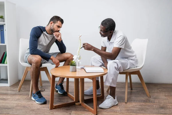 African american rehabilitologist pointing at spine model near thoughtful young man — Foto stock