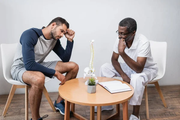 Young man and mature african american doctor looking at spine model near blank notebook — Stock Photo