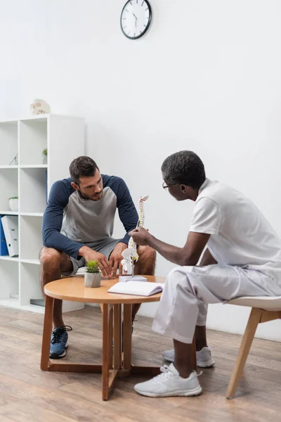 Médecin afro-américain d'âge moyen pointant vers le modèle de colonne vertébrale près du jeune homme dans le centre de réadaptation — Photo de stock