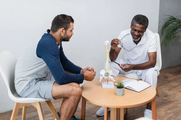 Smiling african american rehabilitologist holding eyeglasses and pointing at spine model near young patient — Fotografia de Stock