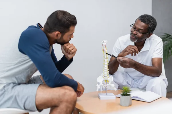 Fisioterapeuta afroamericano apuntando a modelo de columna vertebral cerca de hombre joven en sala de consulta - foto de stock